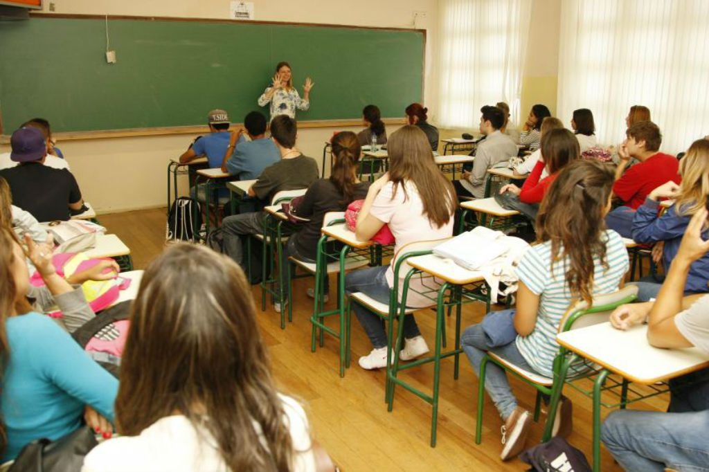 Sala de aula, com alunos sentados em carteiras, e professora dando aula